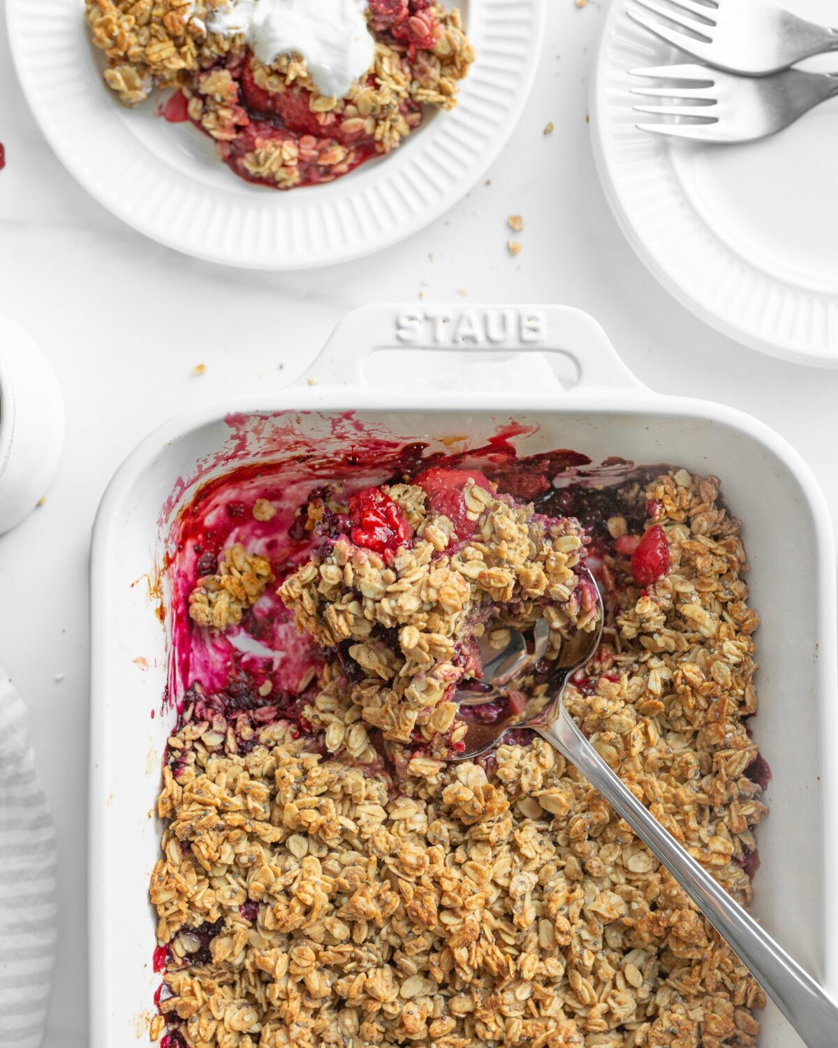 Triple Berry Breakfast Crumble baked in a baking dish. Some of it is scooped onto a small plate and topped with greek yogurt.