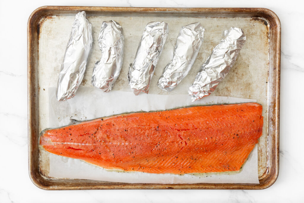 Process shot #1: Large fillet of wild sockeye salmon and foil-wrapped sweet potatoes on a baking sheet.